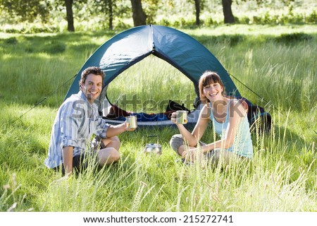Young couple sitting near dome tent on camping trip in woodland clearing, holding mugs, smiling, portrait