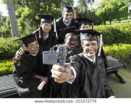 Man filming university students in graduation gowns and mortar boards, using camcorder, smiling