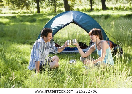 Young couple sitting near dome tent on camping trip in woodland clearing, making celebratory toast with mugs