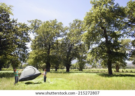 Young couple standing in mid-distance, assembling dome tent on camping trip in woodland clearing