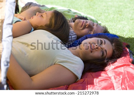 Family lying on sleeping bags in tent entrance on garden lawn, father and children sleeping, mother daydreaming, side view