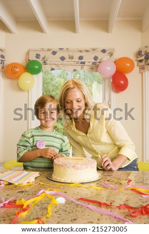 Boy (4-6) sitting at table beside birthday cake, mother holding candles, smiling, front view, portrait