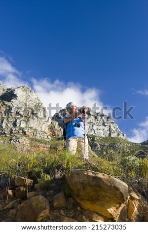 Mature woman hiking on mountain trail, leaning on hiking pole, admiring scenery, smiling, low angle view