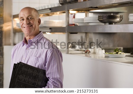 Male restaurant manager standing in commercial kitchen, carrying menus, smiling, side view, portrait