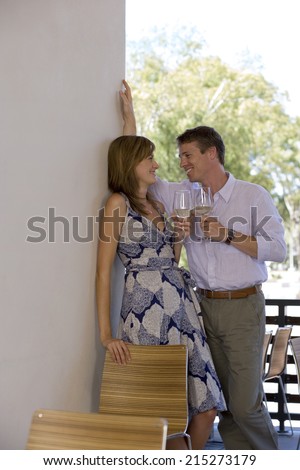 Couple flirting on balcony, holding glasses of white wine, smiling, side view