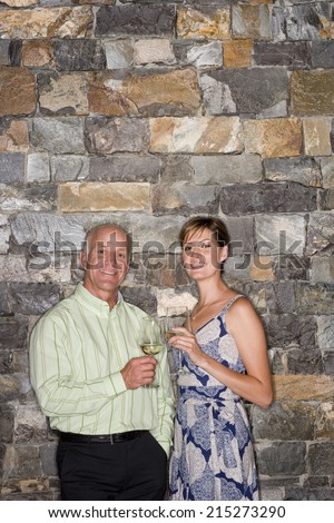 Mature man and young woman standing beside stone wall, holding glasses of white wine, smiling, portrait