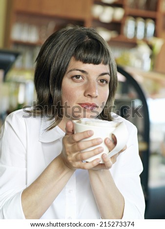 Young woman sitting at cafe table, holding mug of coffee, daydreaming, close-up
