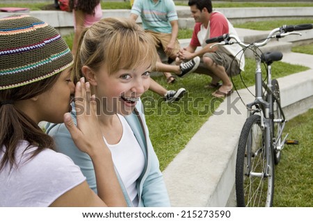 Teenage girl whispering in friend\'s ear, friend smiling, group of teenagers in background