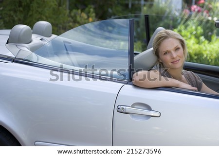 Woman sitting in driving seat of parked convertible car on driveway, smiling, side view, portrait