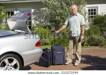 Senior man unloading suitcases on wheels from parked car boot on driveway, smiling, portrait