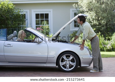 Man standing beside parked convertible car on driveway, checking engine, woman sitting in driving seat, smiling, side view