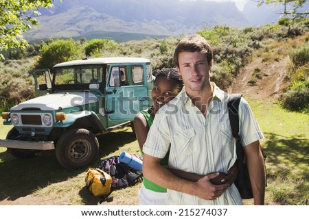 Young couple standing near parked jeep at start of camping holiday, woman embracing man, smiling, portrait