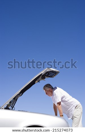 Senior man experiencing car trouble, looking at engine, bonnet raised against clear blue sky, profile