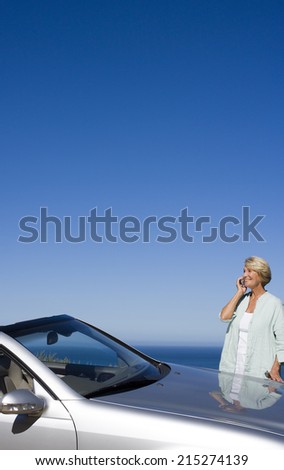 Senior woman standing beside parked convertible car on clifftop overlooking Atlantic Ocean, using mobile phone, smiling, side view