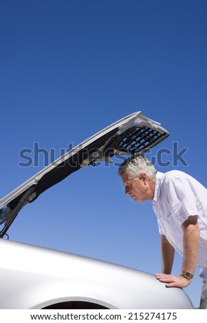 Senior man experiencing car trouble, looking at engine, bonnet raised against clear blue sky, profile