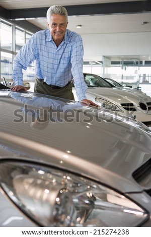Senior man standing in large car showroom, leaning against bonnet of new silver car, smiling, portrait