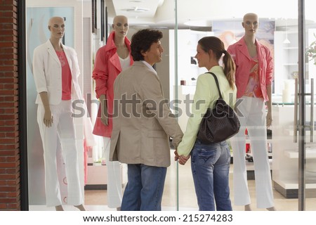 Couple standing outside clothes shop in front of window display, holding hands, smiling, rear view