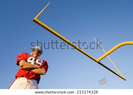 American football player, in red football strip and protective helmet, standing below goal post, arms folded, portrait, low angle view