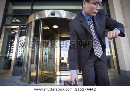 Businessman standing in front of revolving door, checking time on wristwatch, picking up briefcase