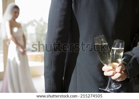 Groom holding two champagne flutes behind back, bride standing in background, focus on groom in foreground, rear view