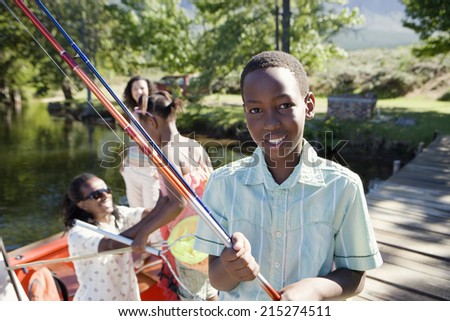 Family standing in motorboat, focus on boy (8-10) standing on lake jetty, holding fishing rod, smiling, portrait