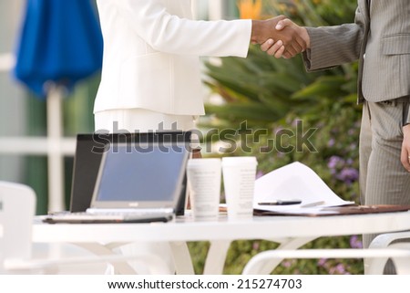 Two businesswomen shaking hands beside pavement cafe table, side view, mid-section