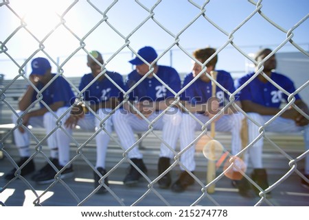 Baseball team sitting on bench in stand during competitive baseball game, view through wire fence, front view (lens flare)