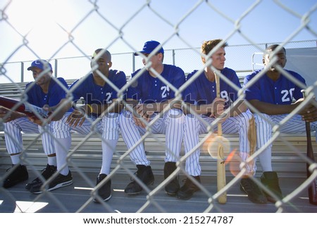 Baseball team sitting on bench in stand during competitive baseball game, view through wire fence, front view (lens flare)