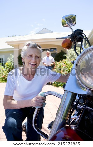 Senior woman polishing motorbike on driveway, crouching down, senior man serving drinks on background, smiling, portrait