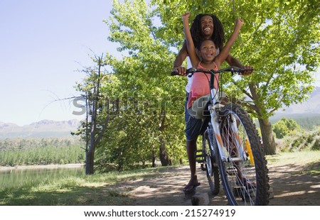 Father and daughter sitting on mountain bike on lakeside woodland trail, girl with arms raised, low angle view