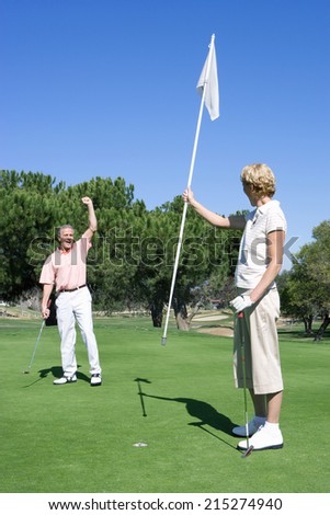 Mature couple playing golf, man punching air in delight at successful putt, woman holding flag on putting green