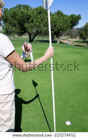 Mature couple playing golf, man lining up golf shot on putting green, woman holding flag