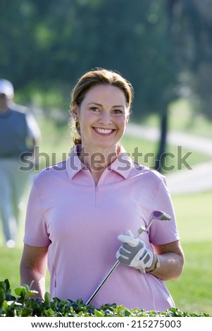 Mature woman in pink polo shirt and golf glove standing on golf course, holding putter, smiling, portrait