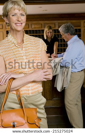 Mature man checking in at hotel reception desk, female receptionist looking on, focus on mature woman standing in foyer in foreground, smiling, portrait