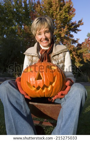 Woman sitting in wheelbarrow in autumn garden, holding Jack O\'Lantern pumpkin, smiling, portrait