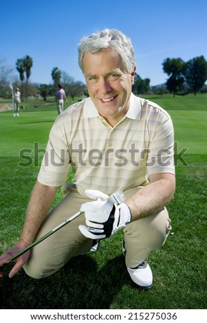 Mature man in striped polo shirt and golf club kneeling on golf course, holding golf club, smiling, front view, portrait