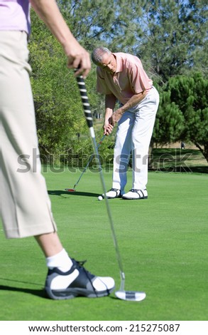 Mature couple standing on putting green, man playing shot, woman watching in foreground