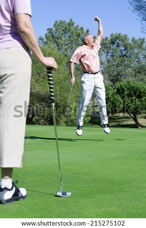 Mature couple playing golf, man punching air in delight at successful putt, woman watching in foreground