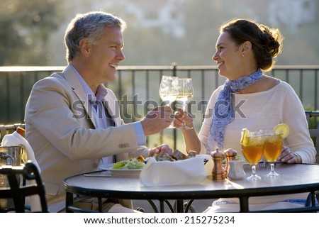 Mature couple dining at outdoor restaurant table, making celebratory toast with wine glasses, smiling, side view