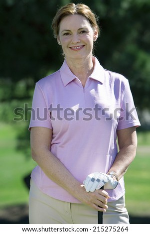 Mature woman, in pink polo shirt and golf glove, standing on golf course, leaning on golf club, smiling, front view, portrait