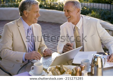 Two mature businessmen sitting at outdoor restaurant table, one man using laptop, smiling, side view
