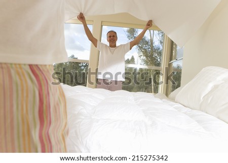 Mature couple making bed, focus on man holding bed sheet in background, smiling