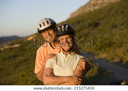 Mature couple, in cycling helmets, standing on mountain trail, man embracing woman, smiling, portrait