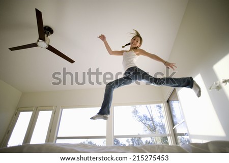 Girl (11-13) jumping up and down on bed, mirroring shape of electric ceiling fan, smiling, portrait, low angle view