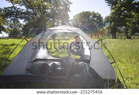 Young man crouching behind incomplete dome tent on camping trip in woodland clearing, holding tent poles, smiling, portrait (lens flare)