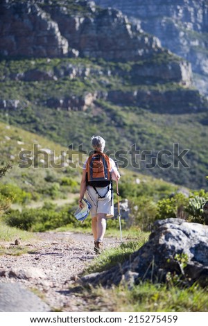 Mature woman hiking on mountain trail, carrying rucksack, using hiking pole, rear view