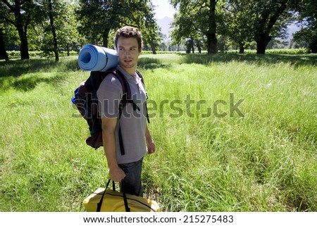 Young man standing in woodland clearing on camping trip, carrying rucksack, tent bag and sleeping bag, smiling, side view