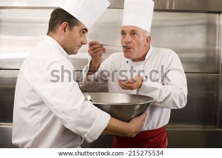 Chef holding large bowl in commercial kitchen, second chef tasting food sample with large spoon