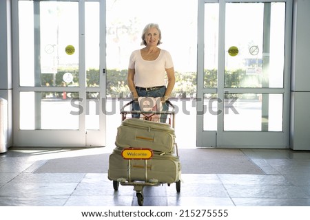 Senior woman entering airport through automatic doors, pushing luggage trolley, smiling, front view, portrait