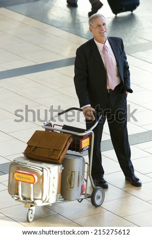 Senior businessman standing with luggage trolley in airport, smiling, elevated view